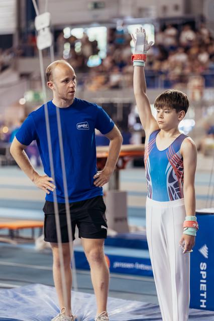 Young male gymnast raises hand to salute before his routine as his coach watches attentively in a packed arena.