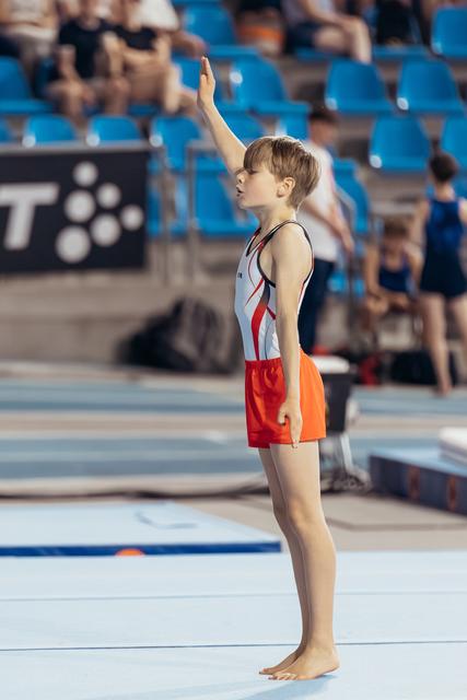 Young male gymnast stands barefoot on floor mat, raising one hand to salute the judges before his routine.