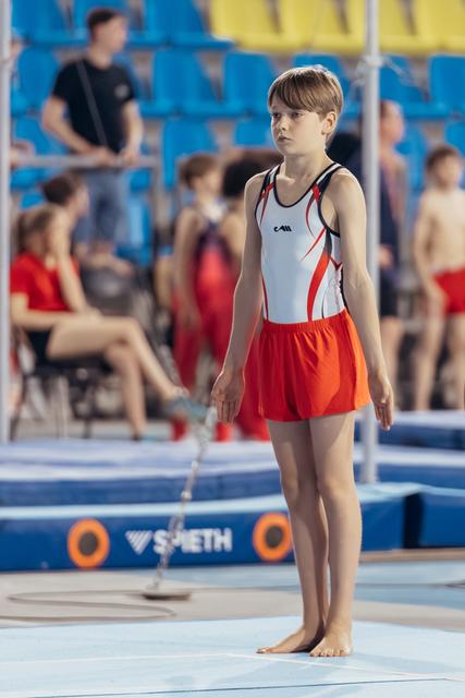 Young gymnast stands barefoot on the mat, composed and focused, wearing white and red leotard with shorts, awaiting their turn.