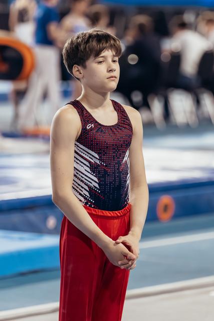 Young male gymnast stands composed, hands clasped, with a calm focused expression before his floor routine.