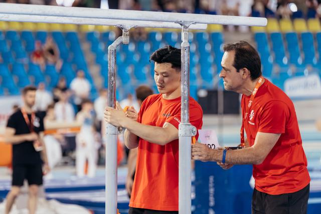 Two coaches in red shirts carefully adjust parallel bars at a gymnastics event, focused and deliberate in their setup.