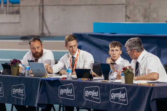 Four male judges in white shirts review scores on laptops at a Gymfed-branded table during a gymnastics competition.