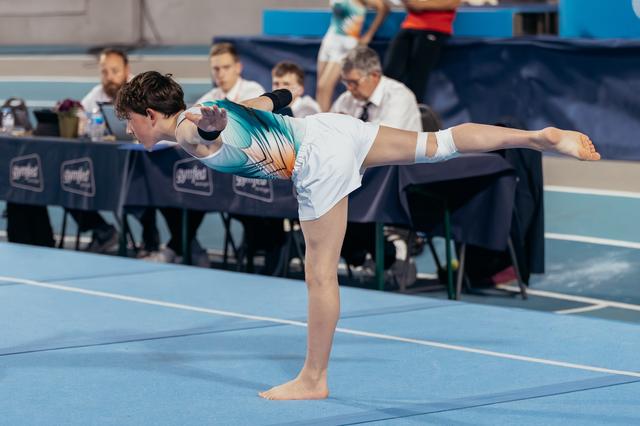 Young gymnast holds a precise arabesque on blue floor, leaning forward with arms extended, focused during competition.