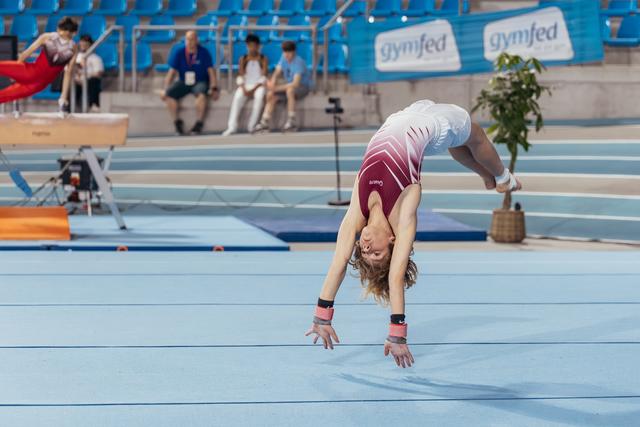 Young gymnast performs a dramatic back walkover on the blue floor, arched deeply with hair flowing, at a Gymfed competition.