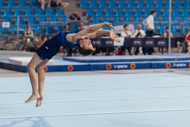 Young gymnast mid-backflip on floor exercise, arms extended, focused expression, at a competitive gymnastics meet.