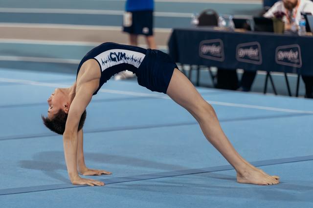 Young gymnast performs a backbend bridge on blue floor mat, head arched back, smiling during competition.