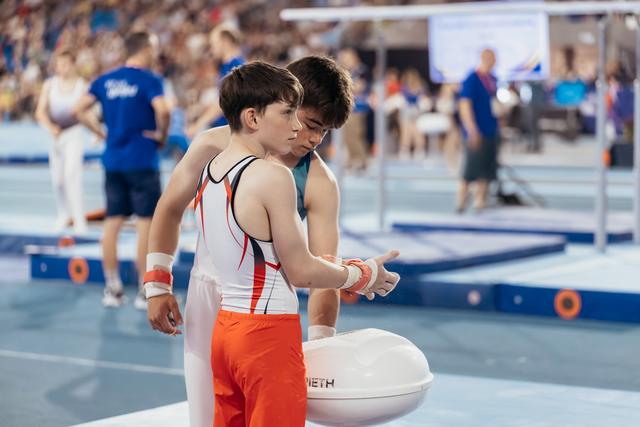 Young male gymnast gets wrist support from coach at chalk bowl, both focused before apparatus routine.