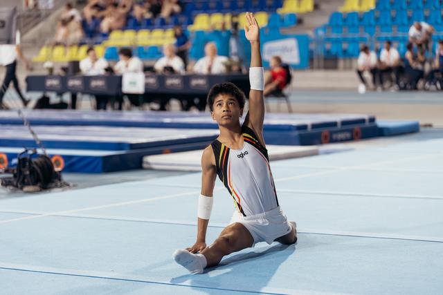 Young male gymnast sits on floor mat, one arm raised high in a composed finishing pose during competition.