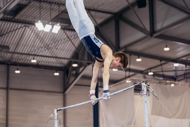 Young male gymnast holds a handstand on the horizontal bar, body fully extended, focused expression in an indoor gym.
