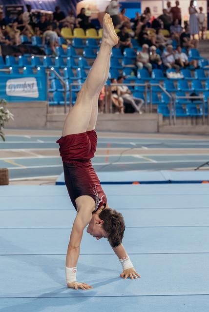Young male gymnast performs a one-armed handstand on the floor, showing intense focus and strength during a competition.
