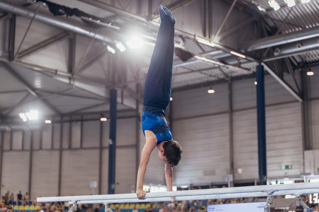 Young male gymnast performs a perfect handstand on parallel bars, body fully extended, in a packed sports arena.