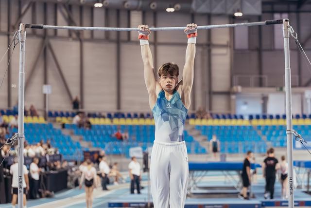 Young male gymnast hangs from the high bar with focused determination, wearing a teal leotard in a large competition arena.