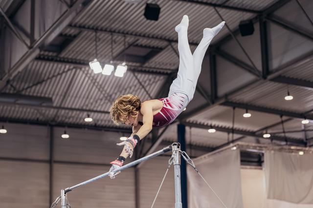 Young male gymnast releases the horizontal bar mid-air, body fully extended upside-down in an indoor gymnastics arena.