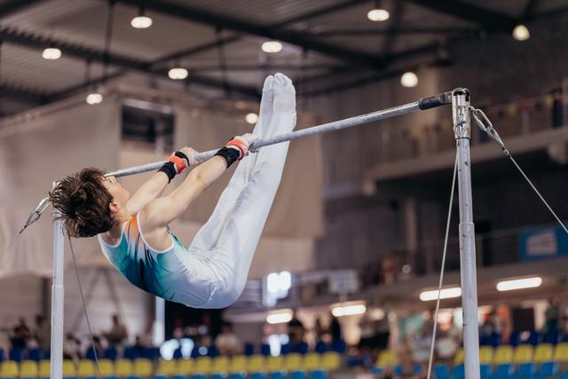 Young male gymnast performs a tuck on the horizontal bar, body curved with intense focus, inside a bright competition hall.