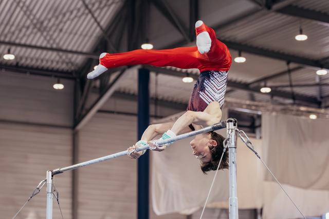 Young gymnast in red performs an inverted release on the horizontal bar, smiling mid-skill in an indoor gymnasium.