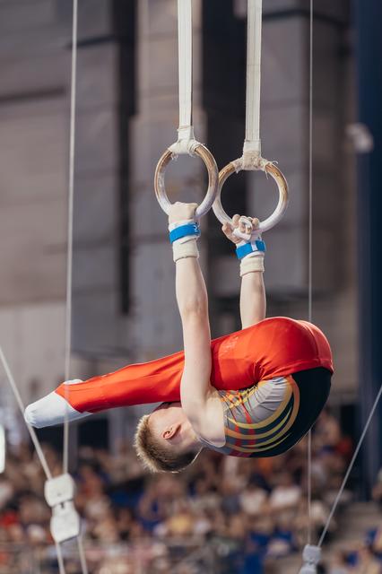 Young male gymnast in red suit performs inverted tuck on still rings, showing intense concentration before a packed crowd.