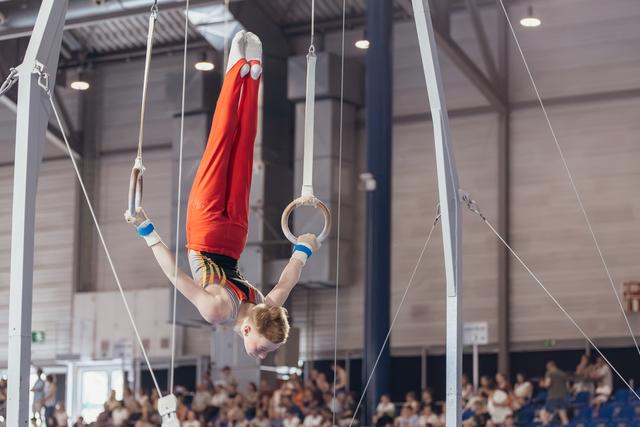 Young male gymnast performs an inverted hold on still rings, body fully extended upside down, wearing red and black leotard.