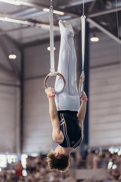 Young male gymnast performs an inverted hang on still rings, wearing white and navy, in a packed indoor arena.