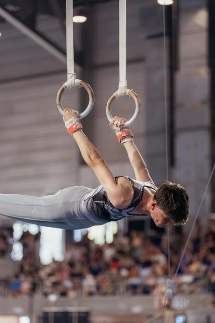 Male gymnast performs an iron cross on still rings, body horizontal, showing remarkable strength and concentration at a gymnastics meet.