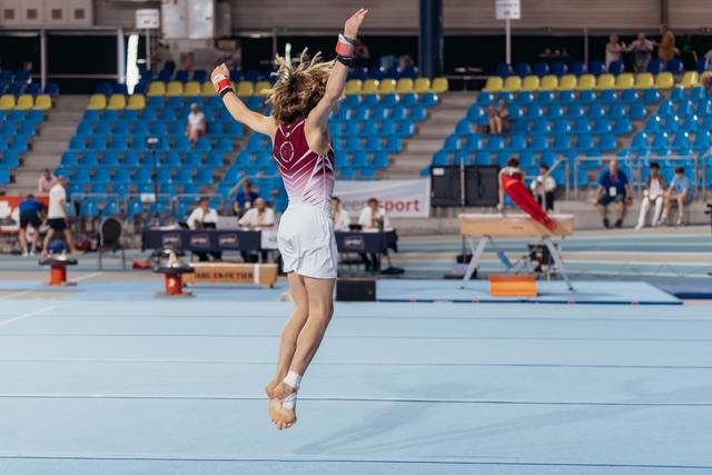 Young gymnast mid-jump on floor exercise, arms raised triumphantly, performing in a large indoor arena with blue seating.