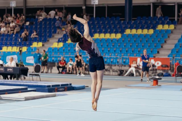 Young gymnast mid-air on floor exercise, one arm raised overhead, poised and focused during indoor competition.