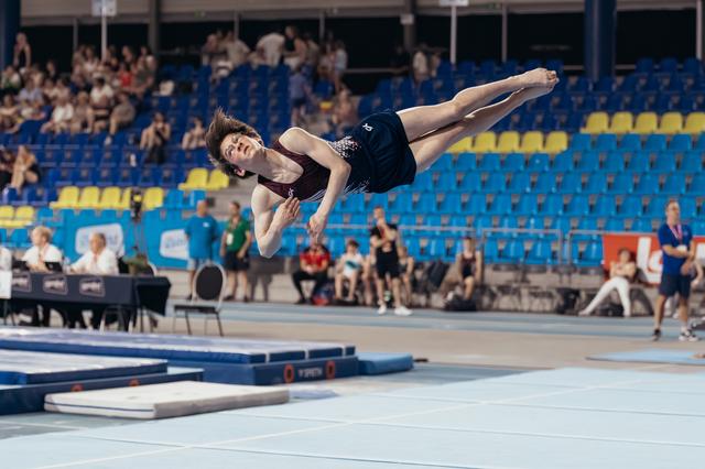 Young male gymnast soaring horizontally during floor exercise, body fully extended, in a packed indoor sports arena.