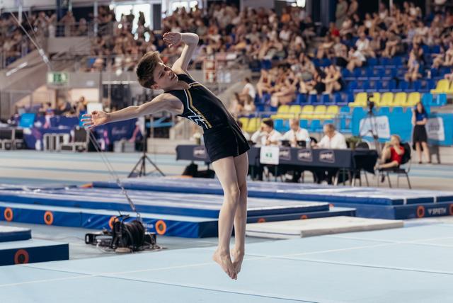 Young male gymnast soars mid-air during floor routine, arms outstretched, focused expression, packed arena in background.