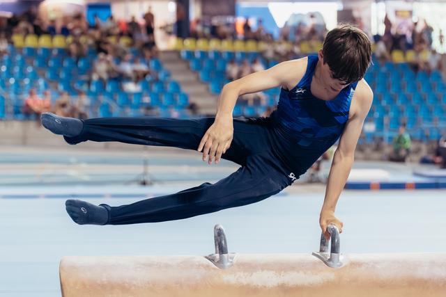 Young male gymnast performs pommel horse routine, legs split mid-air, concentrated expression in arena setting.