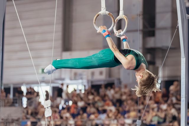 Young male gymnast performs an inverted hold on still rings, body fully extended, intense focus, before a packed crowd.