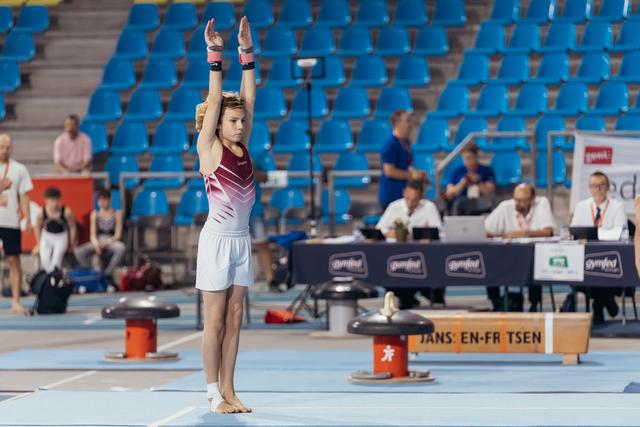 Young gymnast raises arms overhead in a focused salute pose on the floor exercise mat at a Gymfed competition.