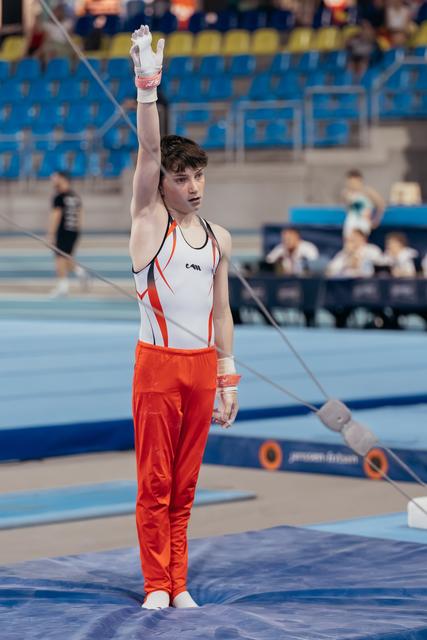 Young male gymnast raises one arm in salute, composed expression, wearing orange and white leotard on competition mat.