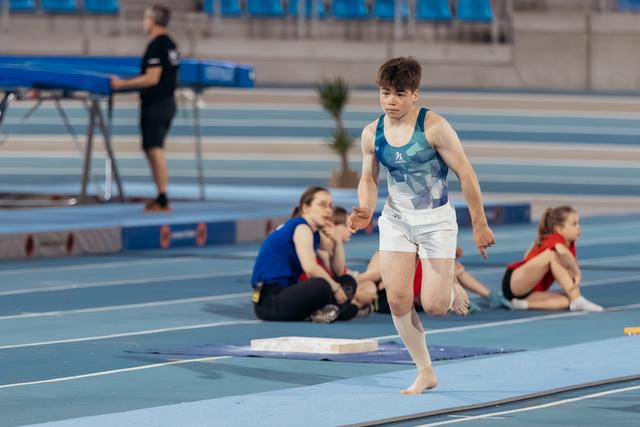 Young male gymnast runs barefoot with focused expression across blue athletics floor, teammates resting in background.