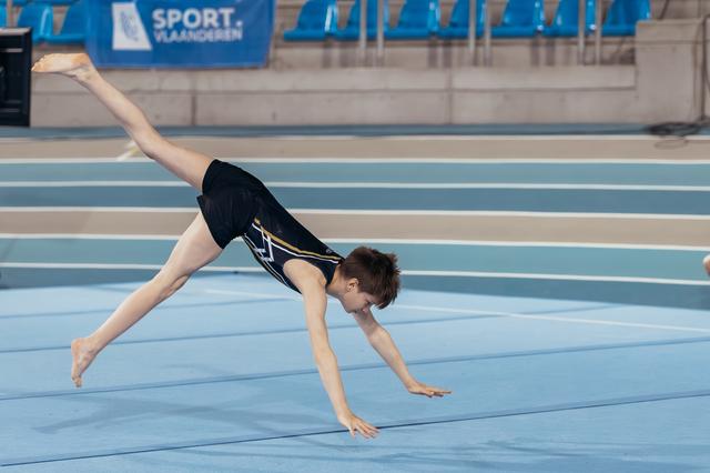 Young gymnast performs a cartwheel on the blue floor mat at a Sport Vlaanderen indoor gymnastics event.