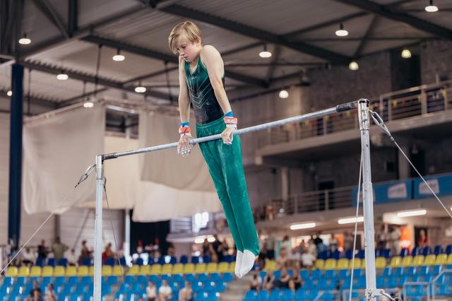 Young male gymnast grips the high bar with focused intensity, body extended, in a packed indoor gymnastics arena.