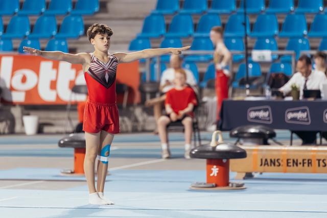 Young male gymnast performing floor exercise, arms outstretched with focused expression, in a competition arena with blue seats.