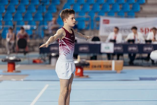 Young male gymnast performs on floor, arms outstretched, focused expression, in a blue-matted arena with judges behind.