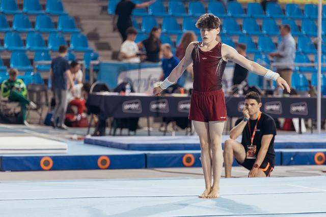 Young male gymnast stands focused on the floor exercise mat, arms extended, barefoot, in a maroon leotard during a competition.