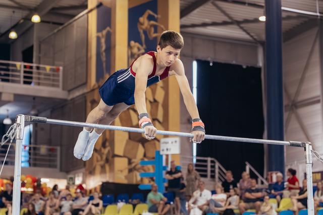 Determined teenage male gymnast grips the high bar mid-routine, body horizontal, intense focus on his face, indoor arena crowd behind.