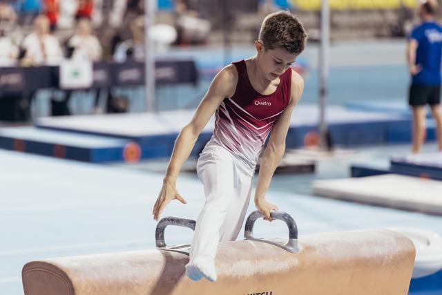 Young male gymnast concentrates intensely while performing on the pommel horse, wearing a red Quatro leotard.
