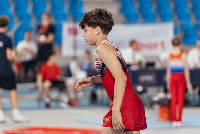 Young male gymnast in red leotard stands focused and composed, preparing for his routine in a busy sports arena.