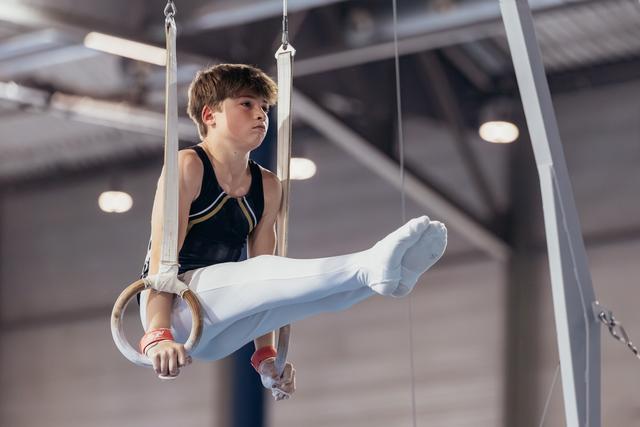 Young male gymnast holds an L-sit on still rings, expression focused and determined, in an indoor gymnastics hall.