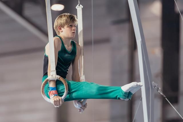 Young male gymnast holds a still rings position with intense focus, legs extended, wearing teal uniform in gymnasium.