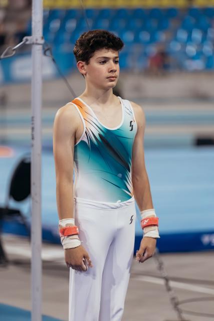 Young male gymnast stands focused and composed beside the high bar, wearing teal leotard with wrist guards, in a competition arena.