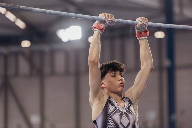 Young male gymnast hanging from the high bar with intense focus, wearing red grips and a geometric leotard indoors.