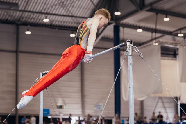Young male gymnast swings powerfully on the horizontal bar, body extended, focused expression in an indoor sports hall.