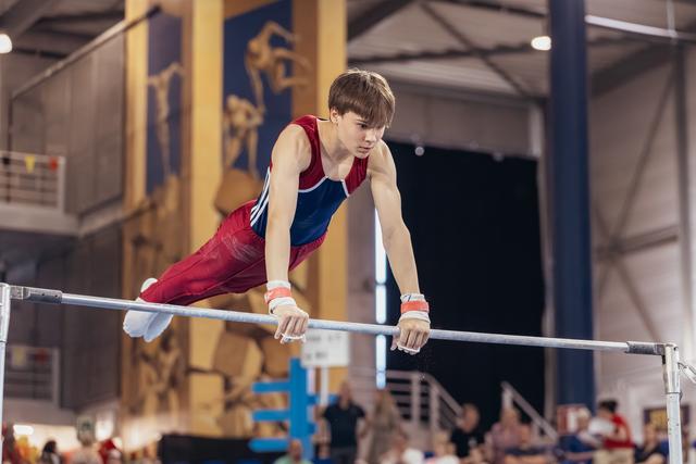 Young male gymnast grips parallel bars with fierce concentration, body horizontal, wearing red and blue leotard indoors.