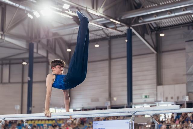 Young male gymnast executes a powerful pike hold on parallel bars, legs raised high, intense focus in an indoor arena.
