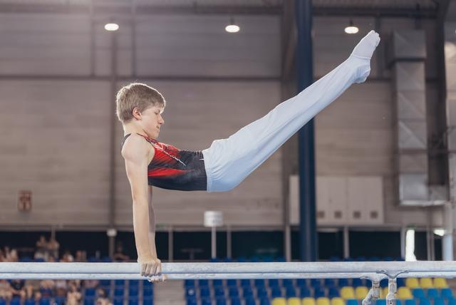 Young male gymnast holds a straddle L-sit on parallel bars, legs raised high, expression intense and focused, in a sports hall.