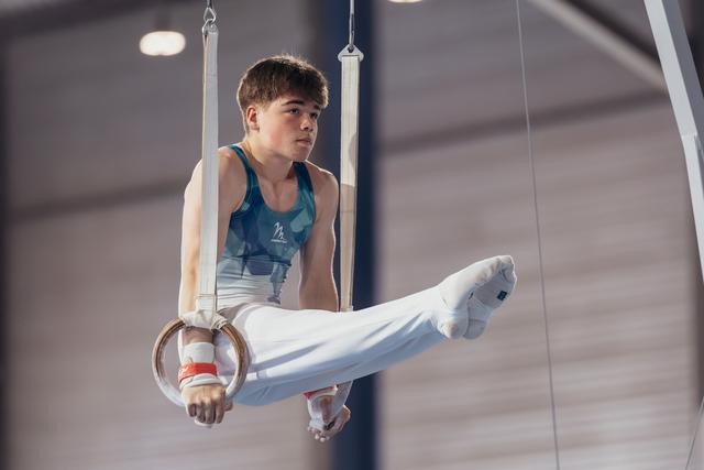 Young male gymnast performs on still rings, legs raised, expression focused and determined in an indoor gymnasium.