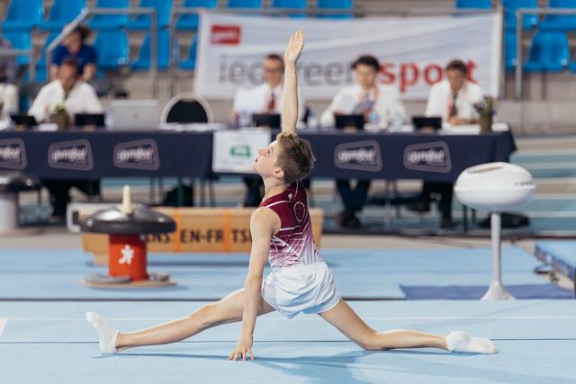 Young male gymnast performs a deep split lunge with arm raised, focused expression, competing on blue floor mat.